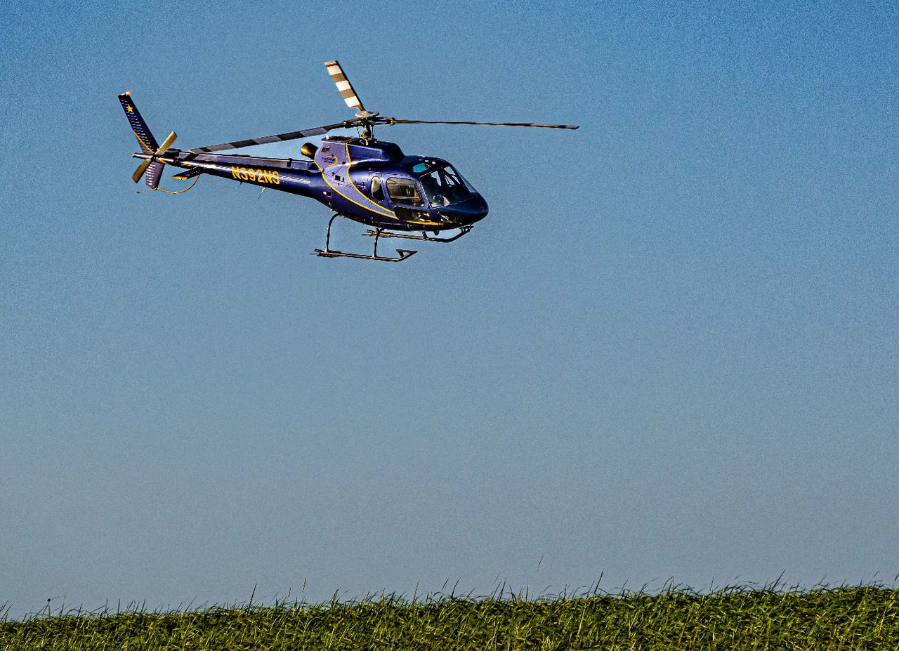 A blue Yakutat Expeditions helicopter in flight low over a green field.